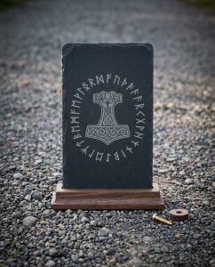 Hand made runic slate plaque engraved with a knotwork Mjolnir inside a full Elder Futhark rune ring, standing in a solid walnut base on grey pebbles.