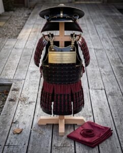 Hand made samurai armor suit in black lacquered steel lamellar with crimson lacing, engraved brass breast plaque and jingasa on a wooden armour stand