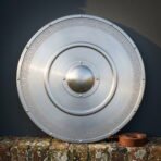 Hand made steel round shield with a domed centre boss, riveted rim and engraved Norse knotwork band leaning on a brick wall beside a leather strap