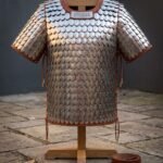 Hand made steel scale armor shirt with brass riveted scales and russet leather binding displayed on a wooden armour stand against a grey stone courtyard