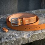 Hand made tan viking leather belt with a stamped Celtic knotwork panel, antiqued roller buckle and brass rivets coiled on a weathered stone ledge