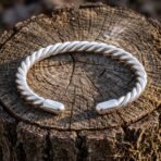 Hand made twisted Viking arm ring in polished silver finish metal with a rope coil body and rounded terminals, resting on a weathered tree stump in sunlight.