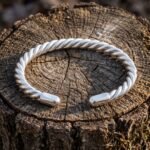 Hand made twisted Viking arm ring in polished silver finish metal with a rope coil body and rounded terminals, resting on a weathered tree stump in sunlight.