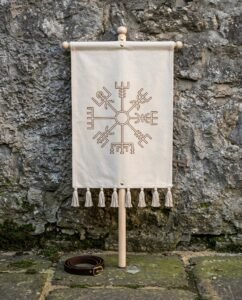 Hand made vegvisir banner in natural cotton canvas hung from a turned ash cross bar with cotton tassels, standing against a weathered stone wall