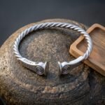 Hand made viking arm ring with a twisted silver finish band, raised spiral terminals and Elder Futhark runes stamped near the ends, resting on weathered grey stone