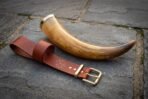 Hand made viking drinking horn with a polished brass rim resting on grey flagstone beside a tan leather belt hanger stamped with a Vegvisir runic compass.