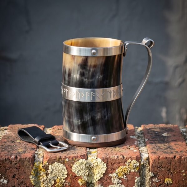 Hand made Viking horn mug with a natural ox horn body, stainless bands, engraved rune band and hardwood base on a brick ledge beside a leather belt hanger