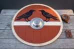 Hand made Viking round shield with twin black ravens on red and white, seen face on with its steel boss and straps