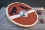 Hand made Viking round shield with twin ravens on red and white and a steel boss, angled on wooden boards with fittings