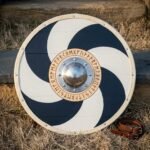 Hand made Viking round shield with a navy and white pinwheel face, riveted rawhide rim, domed steel boss and an engraved Elder Futhark rune ring, leaning on stone.