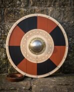 Hand made viking round shield with red and black painted quarters, a carved knotwork ring, domed brass boss and rawhide rim, leaning on a stone wall.