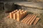 Hand made wooden kubb game in warm beech standing on dry grass with a Vegvisir engraved king, ten kubb blocks, six throwing batons and a green canvas carry bag