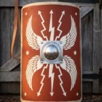 Hand painted roman shield in the curved legionary scutum pattern, red face with white wings and thunderbolts, steel dome boss and rawhide edging against a barn door