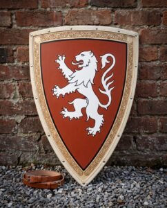 Hand painted wooden heater shield with a white rampant lion on a rust field and knotwork border, leaning on a red brick wall above a leather arm strap