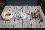 Hand raised steel Viking shield boss with engraved Valknut laid on weathered wood beside leather rim binding, rivets, washers and brown leather arm straps