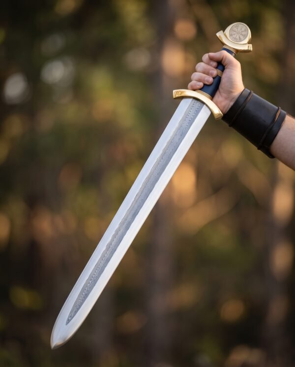 Hand raising a pattern welded Viking sword with blue leather grip and brass hilt against autumn trees