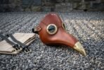 Hand stitched chestnut leather plague doctor mask with brass goggle rims and an engraved brass beak tip, resting on grey gravel beside a stone wall.