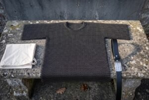 Hand woven butted chainmail shirt in blackened mild steel with short sleeves and a round neck, laid flat across a weathered stone bench outdoors
