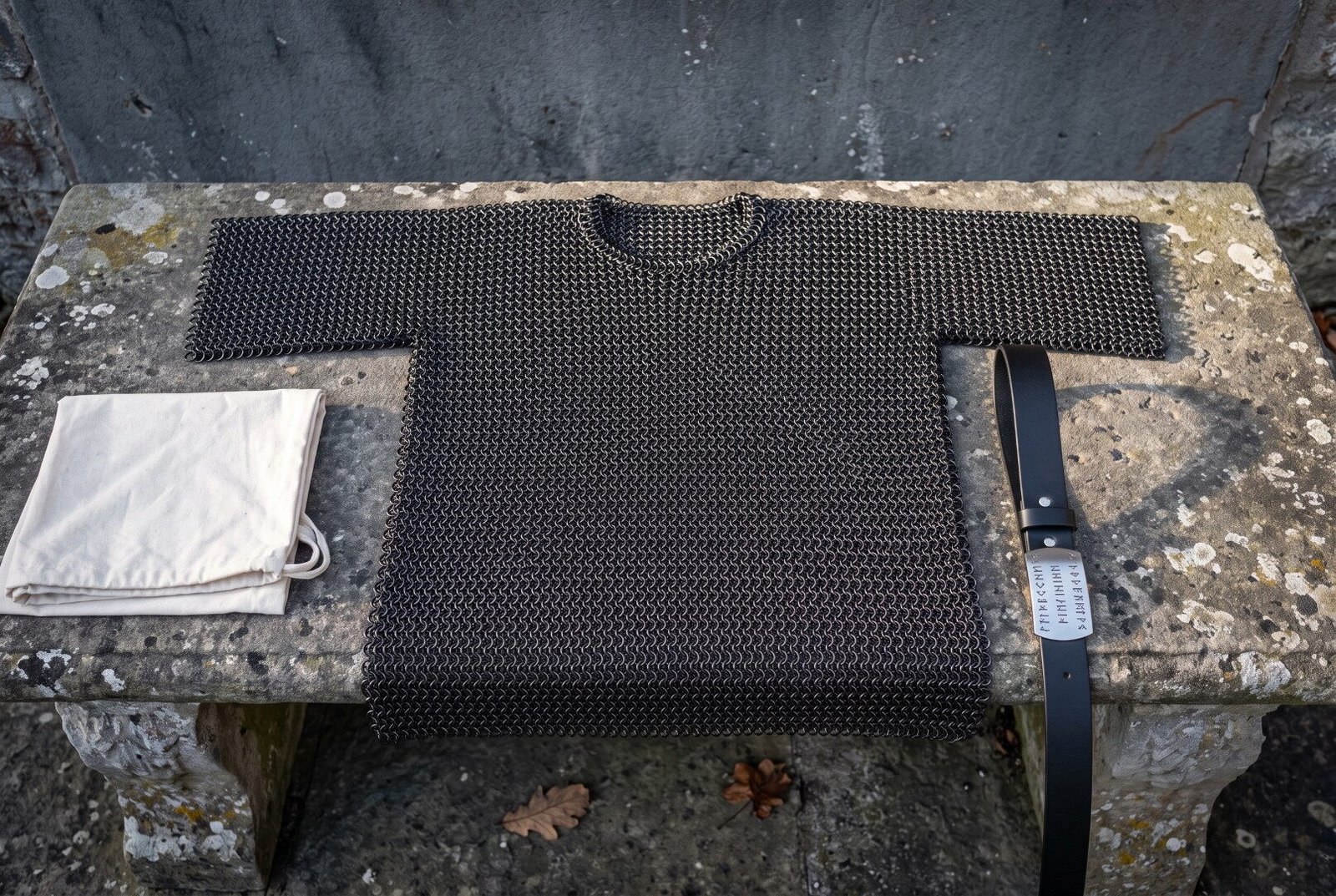 Hand woven butted chainmail shirt in blackened mild steel with short sleeves and a round neck, laid flat across a weathe Hand woven butted chainmail shirt in blackened mild steel with short sleeves and a round neck, laid flat across a weathered stone bench outdoors