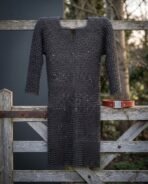 Hand woven chainmail hauberk in blackened mild steel with a four in one butted weave and keyhole neck, hanging on a weathered timber gate in winter light