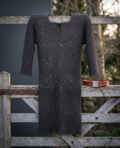 Hand woven chainmail hauberk in blackened mild steel with a four in one butted weave and keyhole neck, hanging on a weathered timber gate in winter light