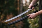 Hands drawing a Damascus katana with black cord handle from its dark wooden saya against autumn trees