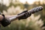 Hardwood baseball bat with barbed wire wrapped barrel and engraved collar held in gloved hands, pale grasses out of focus behind