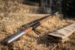 Close up of a hardwood bo staff with a polished steel end cap and black cord grip on dry grass, tan canvas carry bag behind