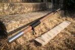 Hardwood bo staff with steel cap and black cord wrap angled across dry grass at the foot of old stone steps, canvas bag alongside