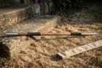 Hardwood bo staff with polished steel end caps, black cord grips and a burned knotwork band lying on dry grass beside stone steps and its canvas carry sleeve