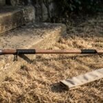 Hardwood bo staff with polished steel end caps, black cord grips and a burned knotwork band lying on dry grass beside stone steps and its canvas carry sleeve
