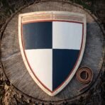 Heater shield in planked wood quartered navy blue and white with a red border and cut knotwork band, lying on a cut tree stump beside a coiled leather strap.