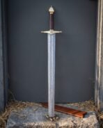 Helm of Awe arming sword with an etched Damascus blade, brass crossguard carrying the eight armed stave, burgundy leather grip and tall brass acorn pommel on stone