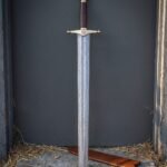 Helm of Awe arming sword with an etched Damascus blade, brass crossguard carrying the eight armed stave, burgundy leather grip and tall brass acorn pommel on stone