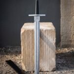 Helm of awe damascus sword with an engraved Aegishjalmur stave below a wide flat guard, black leather grip and bossed wheel pommel standing against a cut stone block