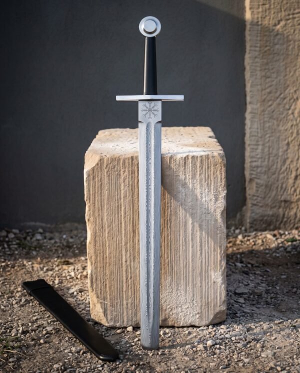 Helm of awe damascus sword with an engraved Aegishjalmur stave below a wide flat guard, black leather grip and bossed wheel pommel standing against a cut stone block