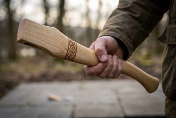 Hickory axe haft with a carved Celtic knotwork band held in one hand outdoors, showing the kerfed eye end
