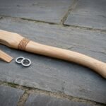 Hickory axe haft with a cut Celtic knotwork band lying on slate paving beside its wooden wedge and two steel ring wedges ready for hanging an axe head