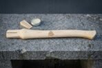 Hickory viking axe handle with a cut Valknut symbol lying on a granite bench beside its wooden wedge and a small tin of finishing wax