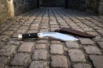 High carbon kukri knife with brass butt cap and black grip, full side view on a cobbled passage before its leather sheath