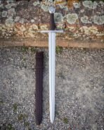 High carbon steel arming sword with wheel pommel, straight cross guard and brown grip laid on gravel beside its dark scabbard