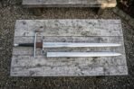 High carbon steel hand and a half sword with ring terminal guard laid on an old wooden table above its polished steel scabbard