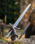 High carbon steel longsword with arched guard and shield pommel raised in a gloved hand above rocks with its scabbard