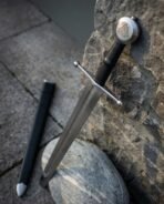 High carbon steel Viking arming sword with Valknut disc pommel leaning on rock beside its black leather scabbard