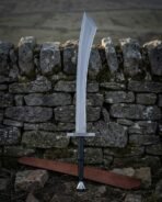 High carbon steel Viking scimitar with broad curved blade and knotwork engraved crossguard standing against a dry stone wall