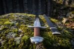 High carbon steel Viking sword seen from the pommel end with brown cord grip on moss next to a brass fitted scabbard