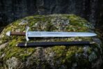 High carbon steel Viking sword with engraved panel above the guard laid on mossy rock over its black and brass scabbard