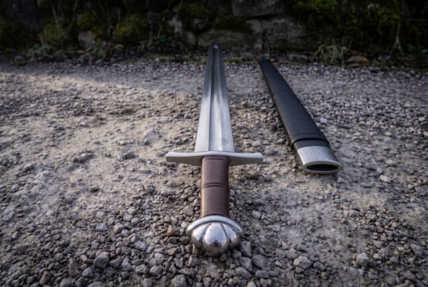 Hilt of a chevron fuller viking sword showing the lobed steel pommel, ribbed brown leather grip and straight cross guard on grey gravel with the black scabbard alongside