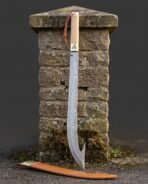 Hooked damascus sabre with a deeply curved pattern welded blade, pale hardwood grip and brass ferrule standing against a dry stone gatepost