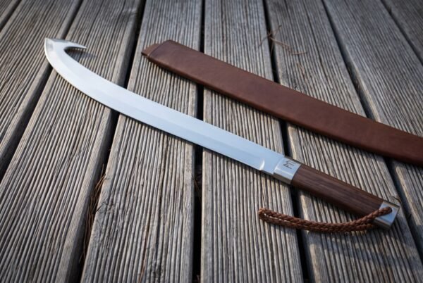 Hooked glaive blade in polished high carbon steel with a dark walnut grip and braided leather lanyard lying on weathered decking beside its brown leather scabbard